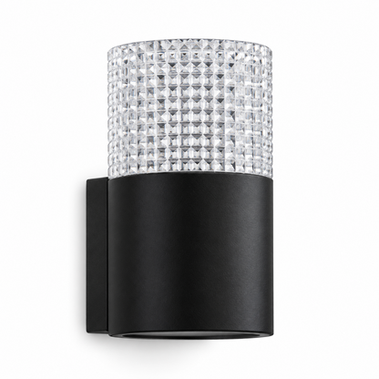 A textured, crystal-effect cylinder shade sits on a sleek black round body with a solid wall bracket, scattering the light into a soft glittering glow after dark.
